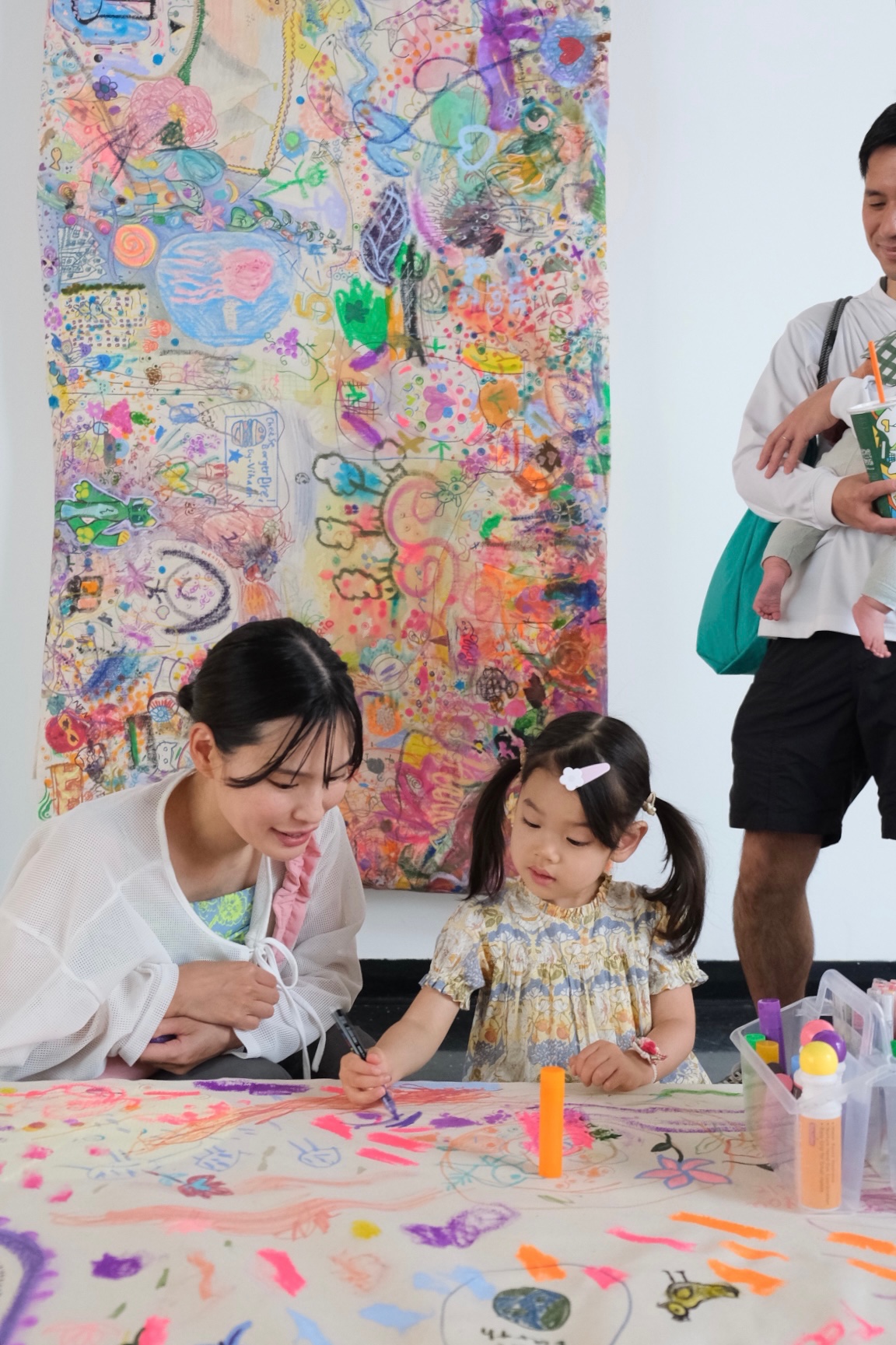 An adult watches closely as a young child adds a mark to the collaborative canvas.