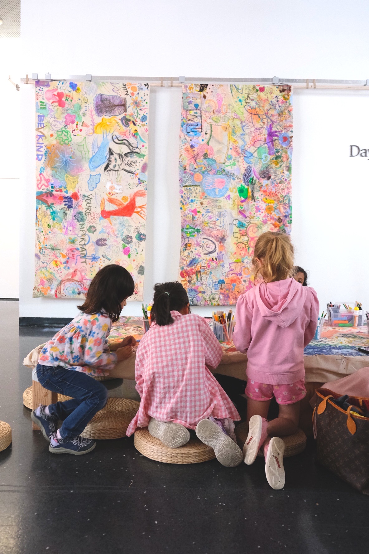 Three children gather around the collaborative canvas, with two finished canvases displayed behind them.