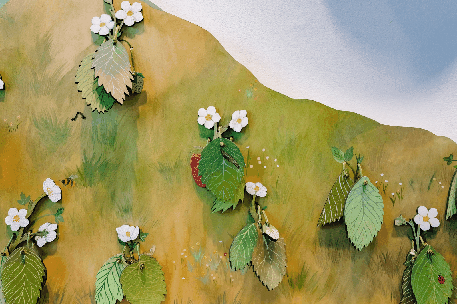 A hand lifts a layered painted leaf to reveal a bright red strawberry beneath it.